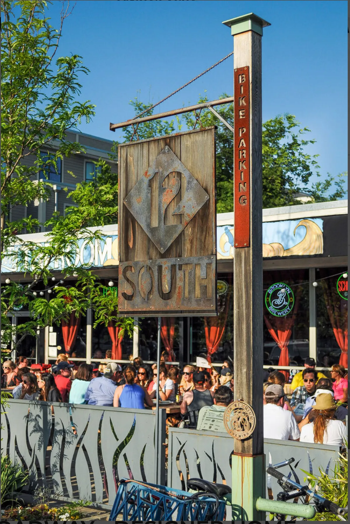 This exterior image shows the lively patio of a local restaurant, with signage, diners, and outdoor seating surrounded by greenery. The scene is bustling with activity, reflecting a vibrant social atmosphere perfect for gathering with friends and enjoying the neighborhood's energy.