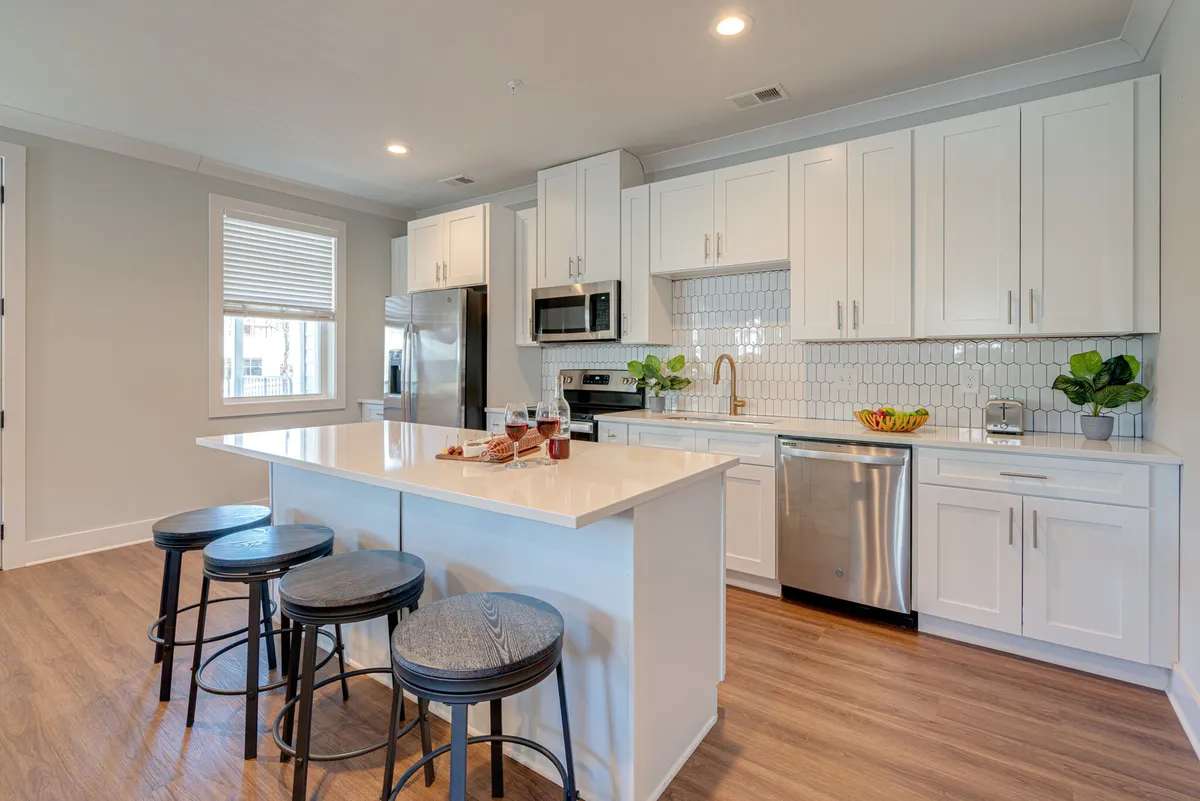 A bright kitchen features a long kitchen island with bar stools, stainless steel appliances including a refrigerator and oven, and sleek white cabinetry. The space is open, social, and set for group meal preparation or casual dining.
