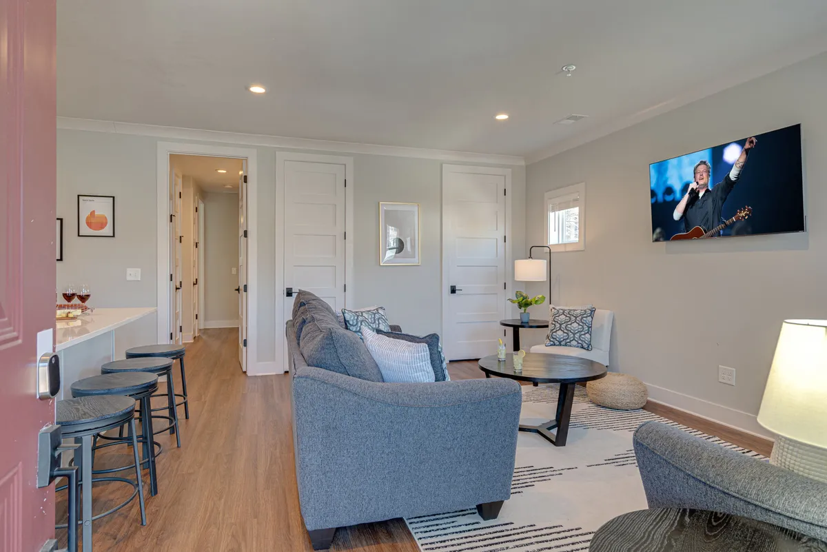 A modern living area with a gray sofa, round coffee table, a wall-mounted TV featuring a performer, and stylish light fixtures. The space feels cozy and intimate, designed for entertainment and group gatherings.