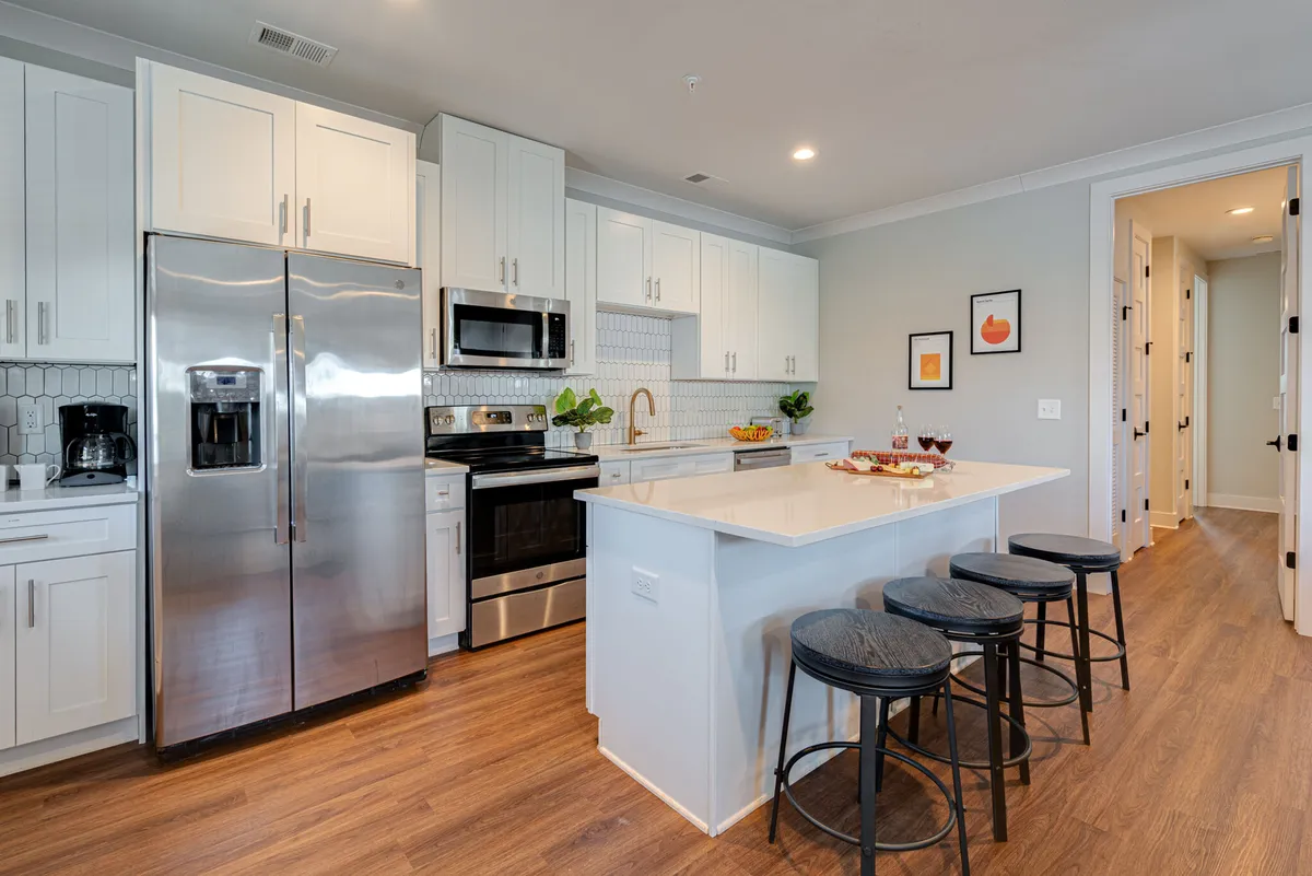 A spacious kitchen features a large kitchen island with seating, a stainless steel refrigerator, white cabinetry, and modern appliances. A decorative fruit bowl adds a touch of warmth to the functional, inviting space. Ample counter space and a clear view of the main cooking area are visible.