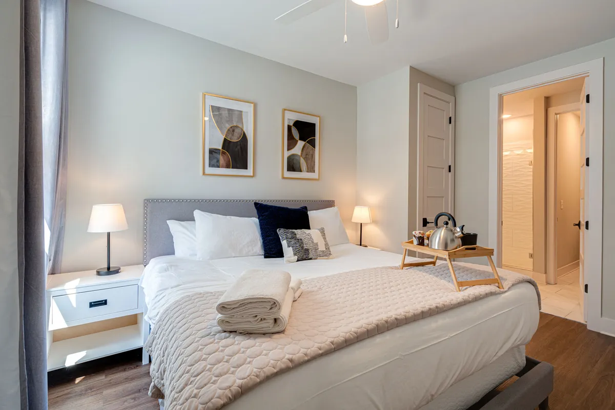 A calm, cozy bedroom scene with a soft bed, decorative pillows, framed artwork, a nightstand with a lamp, and a tray featuring a kettle and cups—suggestive of relaxing mornings or evenings in bed.