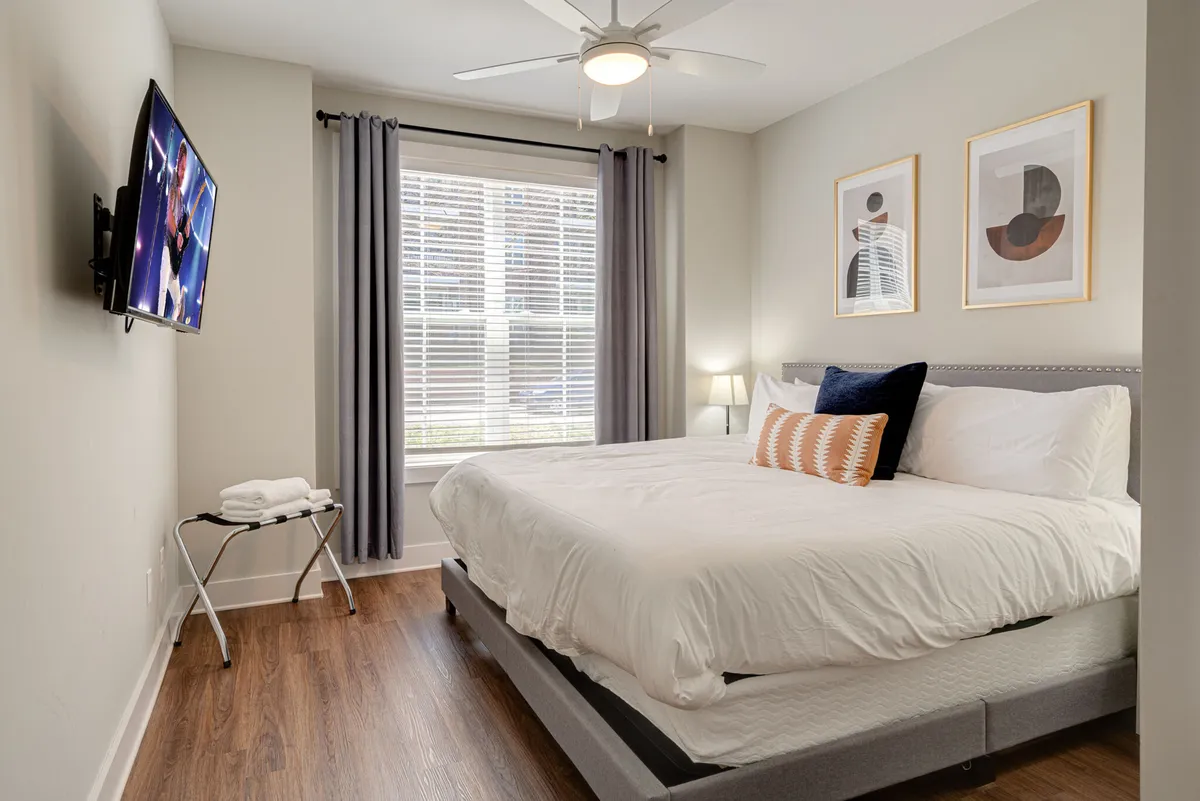 A stylish, cozy bedroom featuring a large bed with crisp white linens, a flat-screen TV mounted on the wall, a window letting in natural light, decorative artwork, and a bedside lamp. The space feels modern and inviting, perfect for relaxing after a busy day.