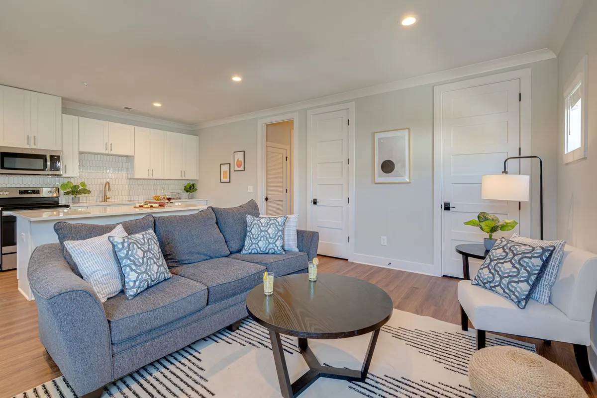An open living room with a comfortable sofa, coffee table, kitchen area in the background, and decorative artwork on the walls. The seamless flow between lounging and dining spaces supports both small groups and large gatherings.