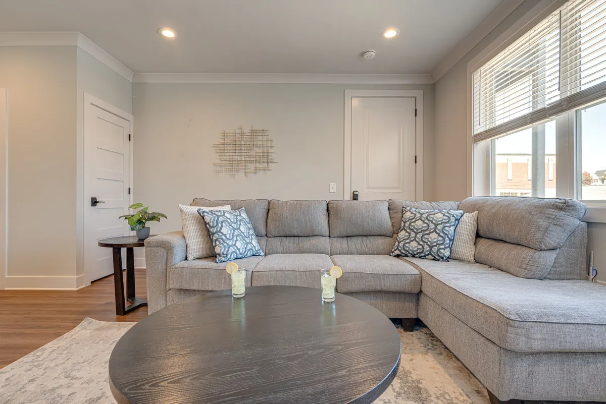 A spacious, open-plan living room featuring a gray sectional sofa adorned with decorative pillows, a round coffee table, and a lush potted plant. The room is bathed in natural light, creating a welcoming, cozy atmosphere ideal for gathering and socializing.
